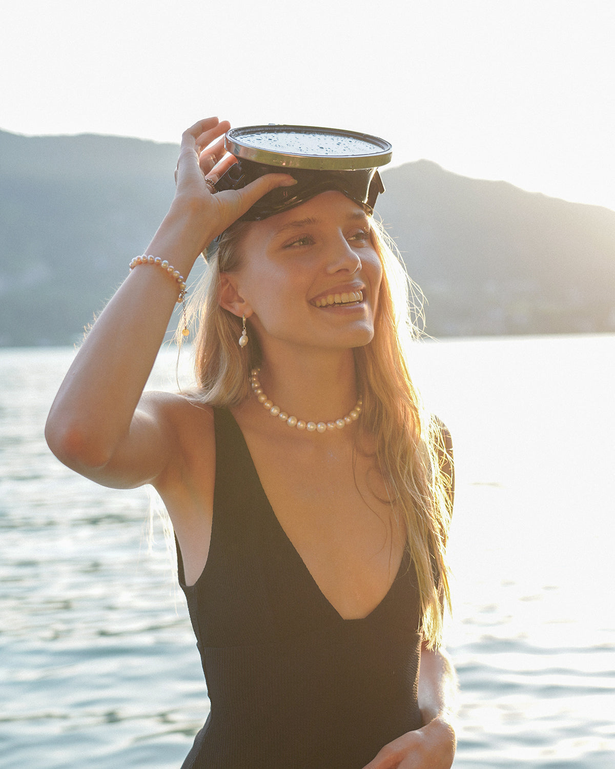 Blonde Frau am See trägt eine Perlenkette, passende Ohrringe und ein Perlenarmband zu einem schwarzen Badeanzug, während sie lächelnd eine Schwimmbrille auf dem Kopf hält; im Hintergrund schimmern Wasser und Berge im warmen Abendlicht.
