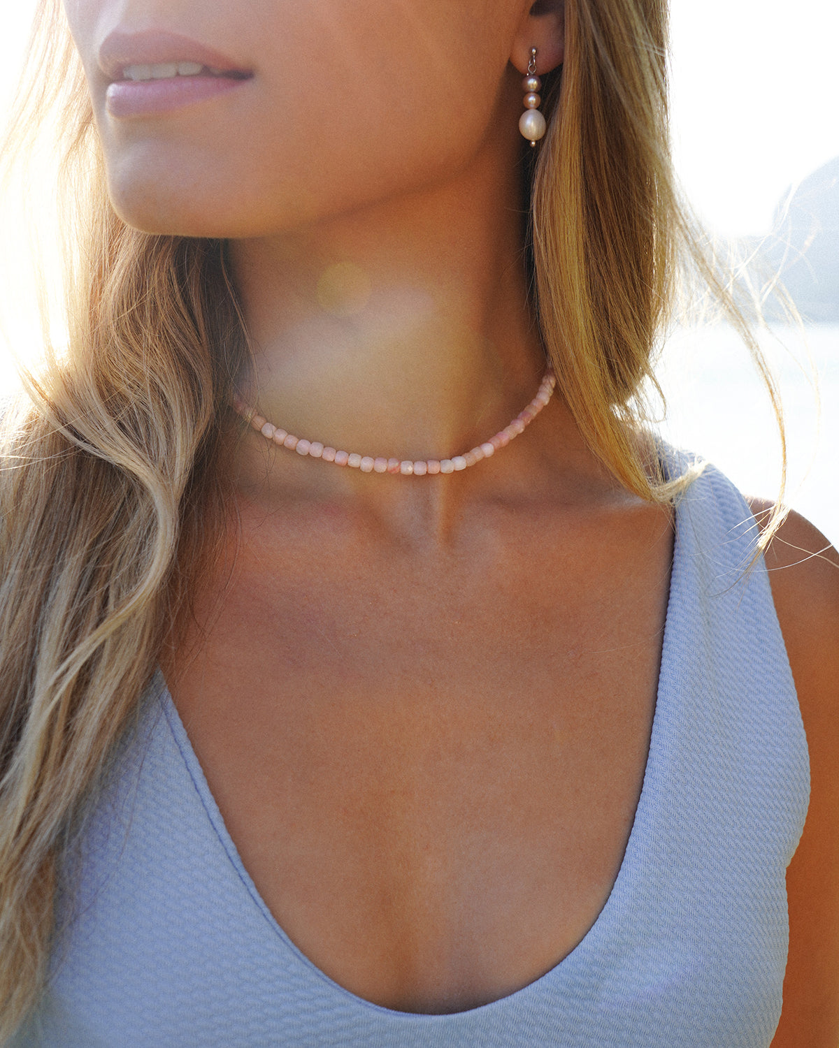 Detailaufnahme einer rosa Opal Halskette als Choker am Hals einer blonden Frau im hellblauen Top, aufgenommen im warmen Sonnenlicht vor See- und Bergkulisse.