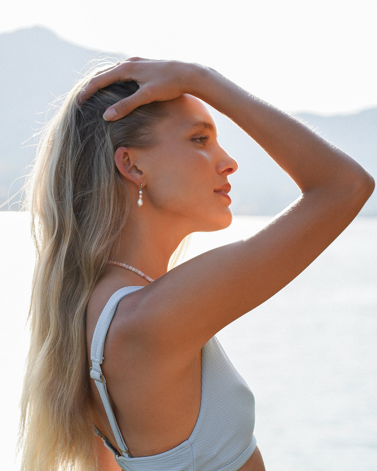 Blonde Frau im hellblauen Kleid trägt dreistufige Perlenohrringe in Rosé- und Weißtönen sowie eine zarte rosa Perlenkette, aufgenommen im warmen Sonnenlicht vor einer See- und Bergkulisse.