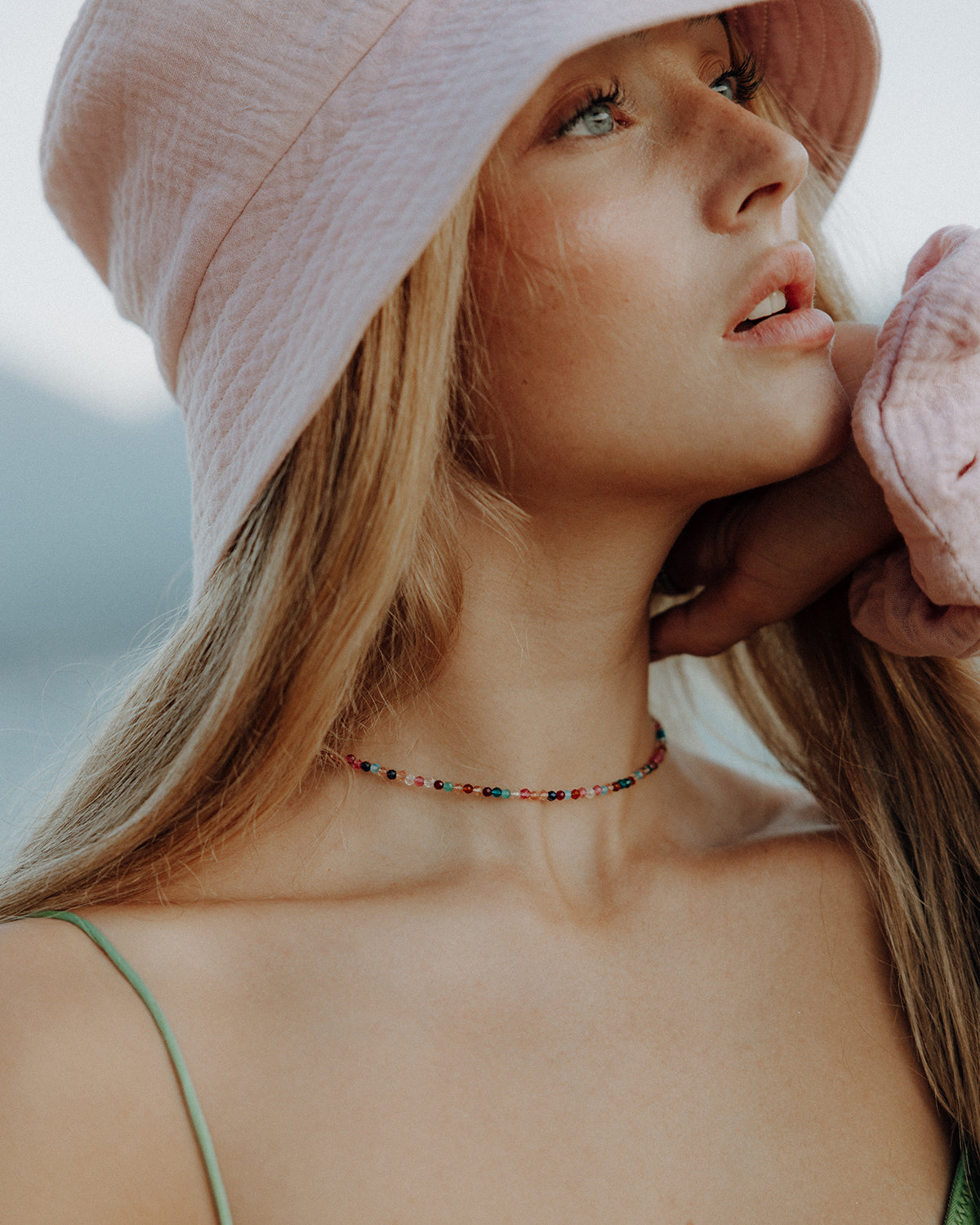 Porträt einer Person mit langem blondem Haar, die einen rosafarbenen Bucket-Hut trägt und leicht nach oben blickt. Sie trägt ein grünes Top und eine feine, bunte Perlenkette. Eine Hand liegt am Nacken, der Hintergrund ist weich und unscharf und erinnert an eine Landschaft.