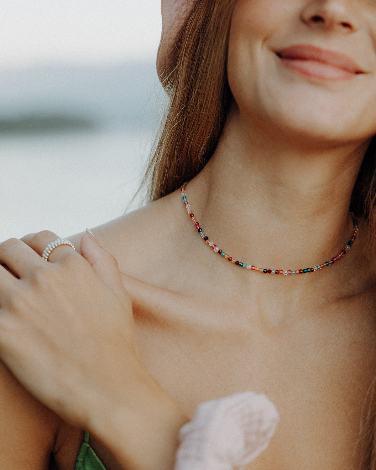 Nahaufnahme einer Person mit leichtem Lächeln, die eine bunte Perlenkette trägt. Eine Hand liegt auf der Schulter, an einem Finger ist ein Perlenring zu sehen. Die Person trägt ein grünes Oberteil und einen hellen Haar-Scrunchie am Handgelenk, der Hintergrund wirkt weich und unscharf.