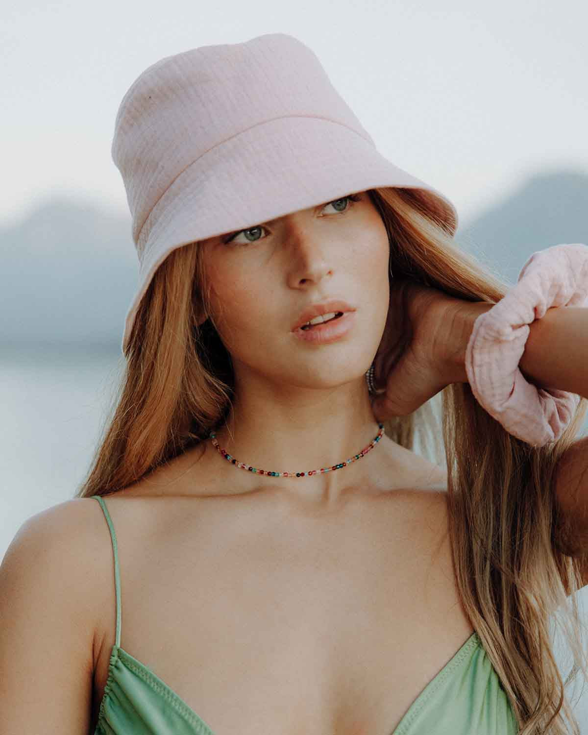 Porträt einer Frau mit langem, hellbraunem Haar, die einen rosa Bucket-Hat trägt und leicht zur Seite blickt. Sie trägt ein grünes Trägertop und eine bunte Perlenkette, während sie eine Hand an ihren Nacken legt. Im Hintergrund sind unscharf Berge und eine helle Landschaft zu sehen.