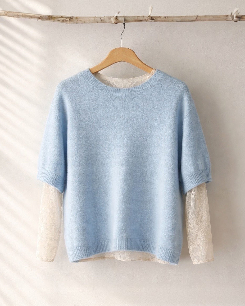 Hellblauer, kurzärmeliger Strickpullover mit Rundhalsausschnitt, der an einem Holzkleiderbügel vor einer hellen Wand hängt. Darunter ist ein langärmliges Spitzentop in Creme sichtbar, dessen Ärmel unter dem Pullover hervorschauen; sanftes Tageslicht wirft dezente Schatten auf die Wand.