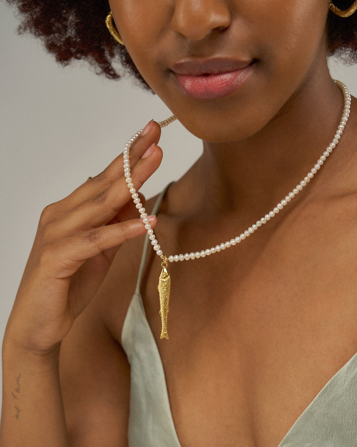 eine Frau die ihre Perlenkette mit goldenem Fischanhänger anhebt und diese präsentiert.
