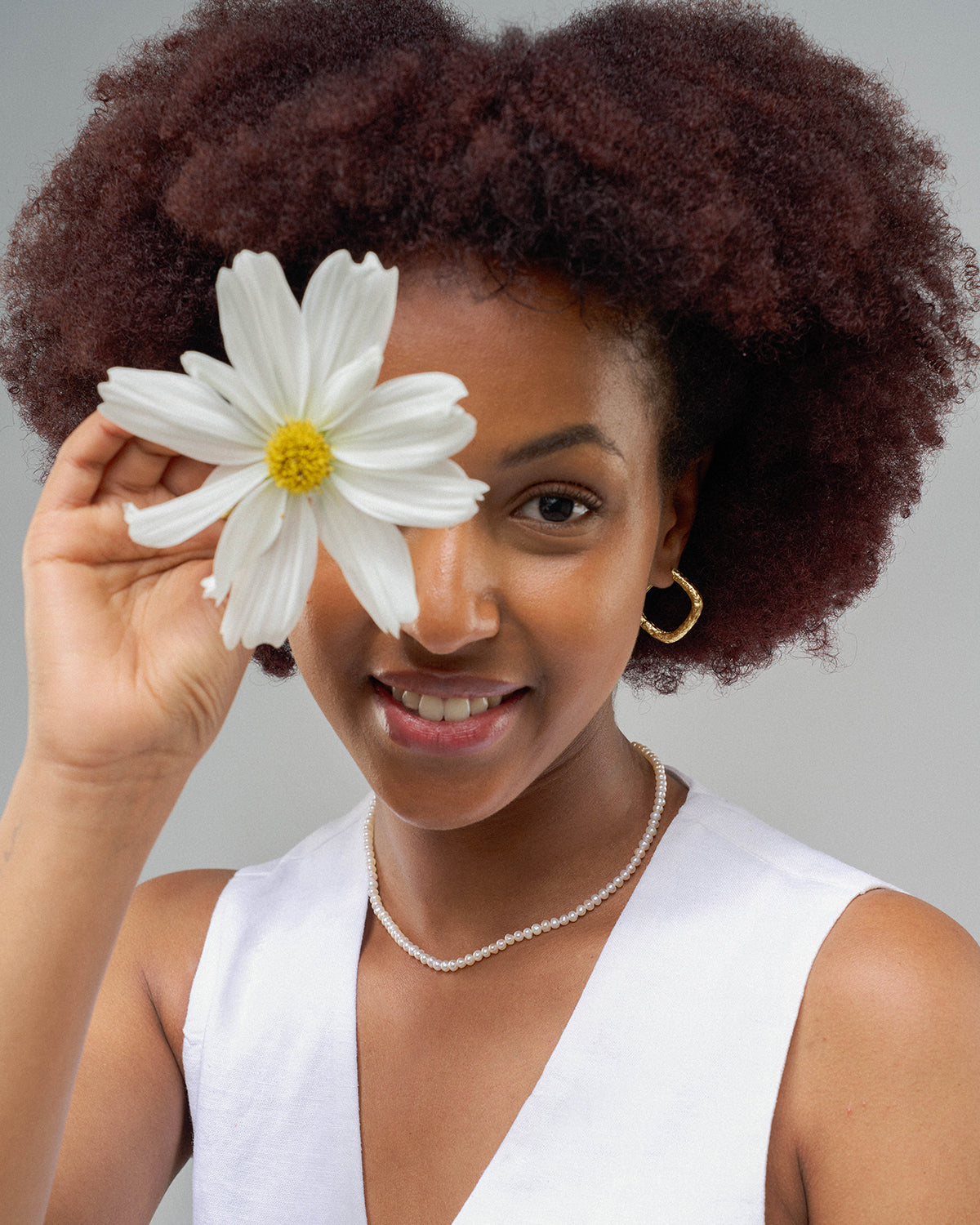 Porträt einer lächelnden Frau mit kurzem, lockigem Haar vor hellem Hintergrund. Sie trägt ein weißes ärmelloses Oberteil, goldene Creolen und eine zarte Perlenkette und hält eine große weiße Blume vor eines ihrer Augen.