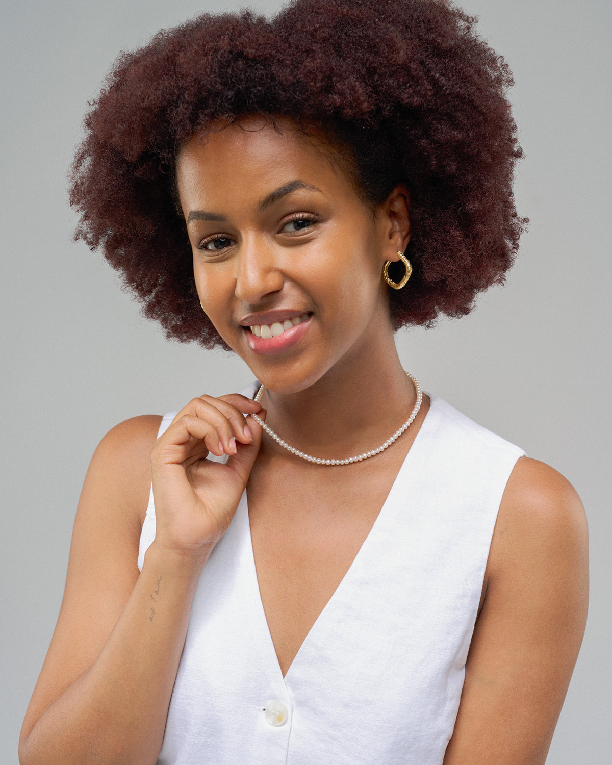Porträt einer lächelnden Frau mit kurzem, lockigem Haar vor hellem Hintergrund. Sie trägt ein weißes ärmelloses Oberteil, goldene Creolen und eine zarte, kurze Perlenkette, die sie mit einer Hand leicht berührt.