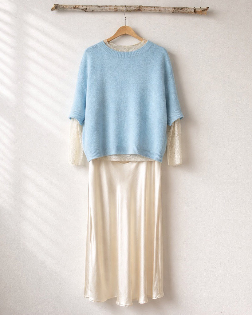 Ein hellblauer, kurzarm Strickpullover hängt auf einem Holzkleiderbügel an einem schlichten Ast vor einer weißen Wand. Darunter ist eine cremefarbene Bluse mit Spitzenärmeln sowie ein langer, fließender Rock in einem sanften Cremeton kombiniert. Weiches Tageslicht fällt von der Seite ein und erzeugt zarte Schatten auf der Wand.