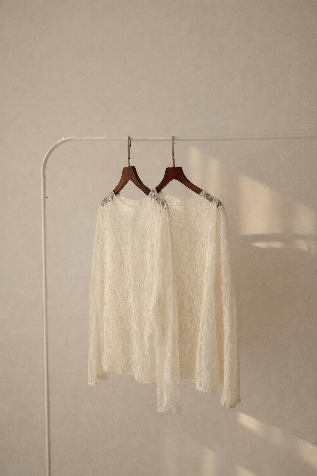 Zwei cremefarbene, langärmelige Spitzenblusen hängen auf dunklen Holzkleiderbügeln an einer schlichten weißen Kleiderstange vor einer hellen Wand. Warmes Sonnenlicht fällt von der Seite ein und wirft weiche Schatten auf die Wand.