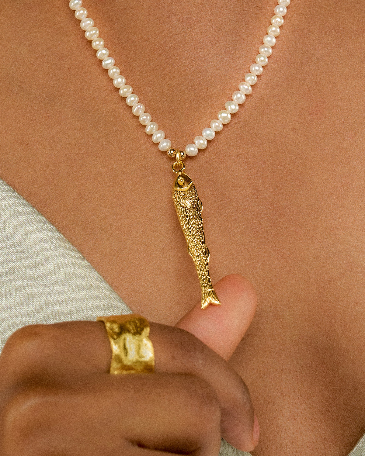 eine Nahaufnahme von einer Perlenkette mit goldenem Fischanhänger. Der Anhänger wird vom Daumen der Frau leicht angehoben.