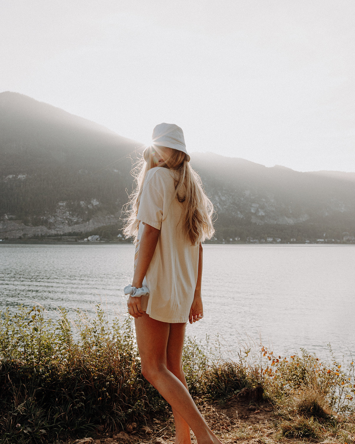 Frau mit langem, blondem Haar trägt einen hellblauen Bucket Hat und einen hellblauen Scrunchie am Handgelenk, steht am Ufer eines Sees mit Berglandschaft im Hintergrund im warmen Gegenlicht der Sonne.
