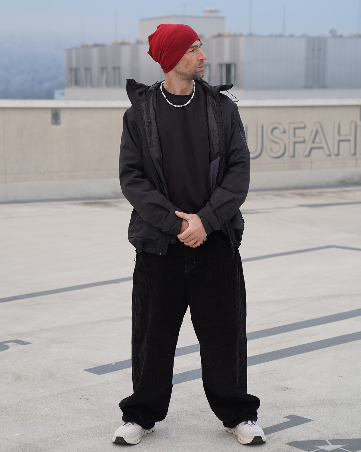 Mann mit roter Mütze und schwarzer Jacke steht auf einem Parkdeck und trägt eine Perlenkette für Herren aus weißen Perlen, moderner urbaner Look mit sportlichem Stil.